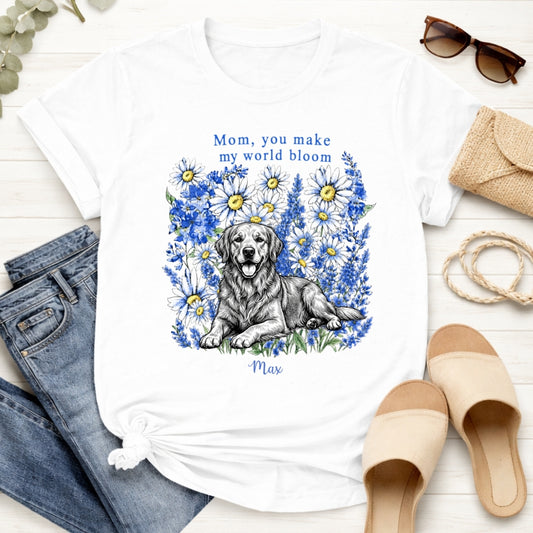 A white custom dogs t-shirt featuring a graphic print of a golden retriever dog surrounded by blue flowers, with the text 'Mom, you make my world bloom' and dog's name written below. The t-shirt is styled in a flat-lay arrangement with blue jeans, beige slide sandals, sunglasses, a burlap clutch, and a rope on a white wood background.