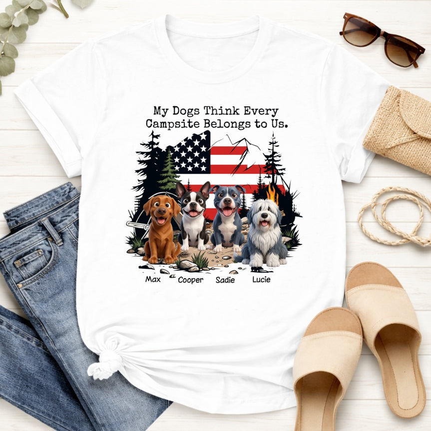 A white graphic t-shirt laid flat on a light wood surface, styled with jeans, sunglasses,... The shirt features a printed design of dogs sitting in front of a campsite scene with an American flag, trees, tent, fire, and the text 'My Dog Thinks Every Campsite Belongs to Us.