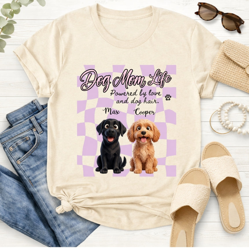 A sand graphic T-shirt laid flat on a light wood surface, styled with jeans, sunglasses, a straw bag, rope, and beige slides. The shirt has a lavender checkerboard graphic with the text 'Dog Mom Life' and 'Powered by love and dog hair.