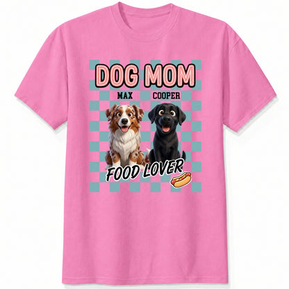 A pink short-sleeve t-shirt with a graphic print featuring custom dogs and the text 'DOG MOM FOOD LOVER' on a light blue checkerboard background.