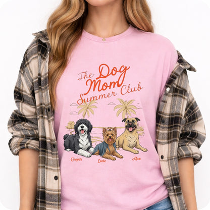 A woman wearing a light pink graphic t-shirt with the text 'The Dog Mom Summer Club' and illustrations of three dogs under palm trees, styled with an open brown and gray plaid flannel shirt over her shoulders.