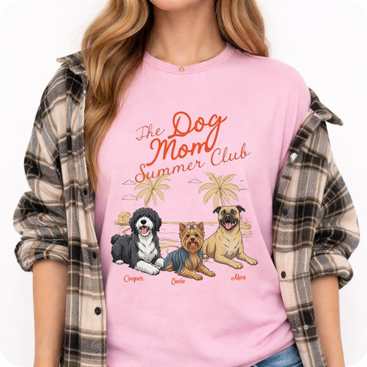 A woman wearing a light pink graphic t-shirt with the text 'The Dog Mom Summer Club' and illustrations of three dogs under palm trees, styled with an open brown and gray plaid flannel shirt over her shoulders.