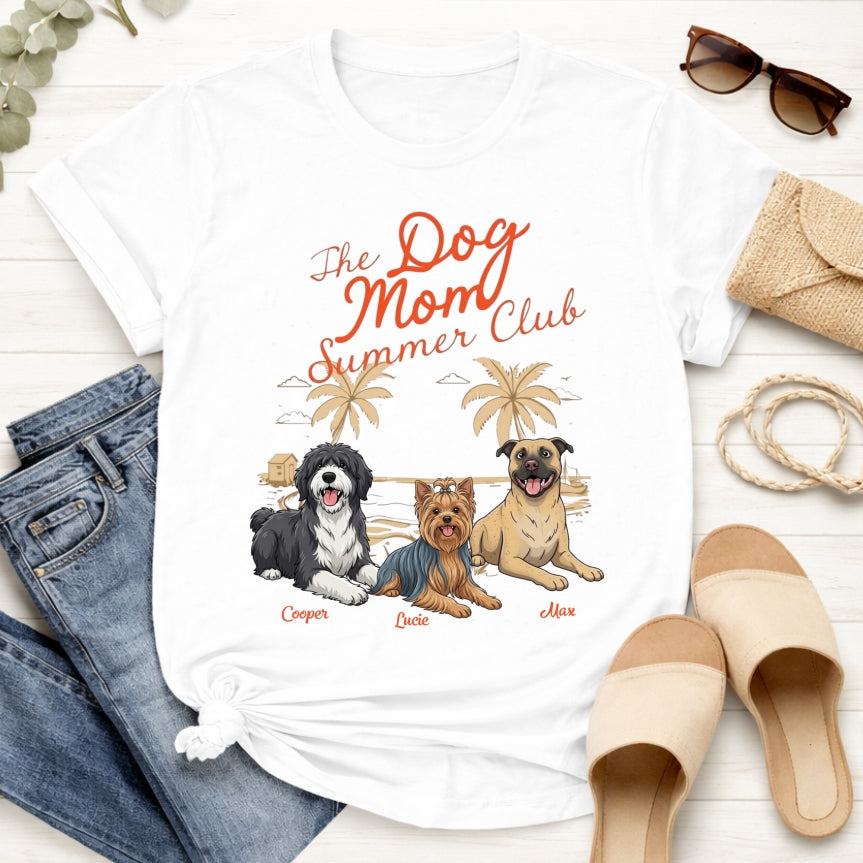 A white graphic T-shirt laid flat on a light wood surface, styled with jeans, sunglasses, a clutch, rope, and slide sandals. The shirt features the text 'The Dog Mom Summer Club' in orange script above an illustration of three dogs with palm trees and the Dog's Names underneath.