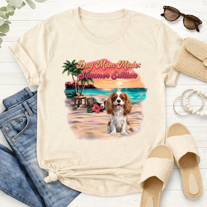 A sand graphic t-shirt featuring a 'Dog Mom Mode: Summer Edition' design with a dog on a beach, styled in a flat lay with jeans, sunglasses, a clutch, and sandals. There are many dog breeds to choose from.