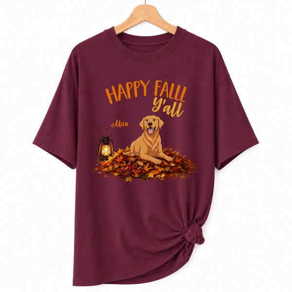 A maroon short-sleeve t-shirt hanging on a wooden hanger against a white background. The shirt features a graphic print of a golden retriever dog sitting on a pile of orange and yellow autumn leaves next to a lantern, with the text 'HAPPY FALL Y'all' and custom dog's name written above the illustration.