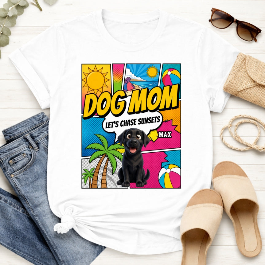 A white-colored graphic t-shirt styled in a flat lay with blue jeans, beige slide sandals, sunglasses, and a woven clutch on a white wood background. The t-shirt features a colorful comic-style graphic on the front with the text 'DOG MOM LET'S CHASE SUNSETS' and custom dogs art and dog's names with palm trees, a beach ball, and a sun.