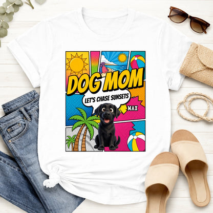 A white-colored graphic t-shirt styled in a flat lay with blue jeans, beige slide sandals, sunglasses, and a woven clutch on a white wood background. The t-shirt features a colorful comic-style graphic on the front with the text 'DOG MOM LET'S CHASE SUNSETS' and custom dogs art and dog's names with palm trees, a beach ball, and a sun.