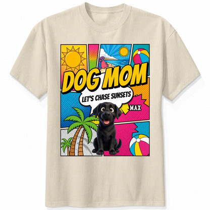 A sand short-sleeve t-shirt featuring a colorful comic-style graphic on the front with the text 'DOG MOM LET'S CHASE SUNSETS' and custom dogs art and dog's names with palm trees, a beach ball, and a sun.