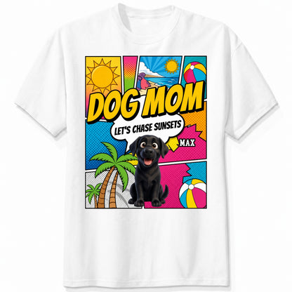 A white short-sleeve t-shirt featuring a colorful comic-style graphic on the front with the text 'DOG MOM LET'S CHASE SUNSETS' and custom dogs art and dog's names with palm trees, a beach ball, and a sun.