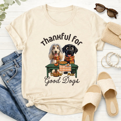A sand-colored graphic t-shirt styled in a flat lay with blue jeans. The t-shirt features a graphic of custom dogs sitting at a table with the text 'Thankful for Good Dogs' and custom with dog's names