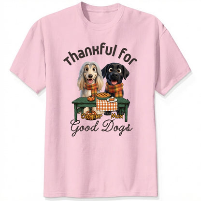 A pink short-sleeve crewneck t-shirt featuring a graphic print of custom dogs sitting at a table with autumn food, with the text 'Thankful for Good Dogs' and custom dog's names written around the illustration.