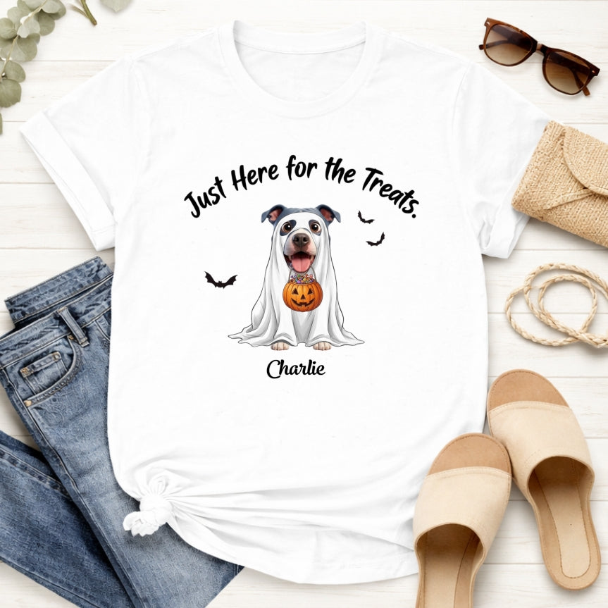 A white graphic T-shirt laid flat on a light wood surface, styled with jeans, sunglasses,...The shirt has a printed dog dressed in a white ghost holding a jack-o'-lantern, with the text 'Just Here for the Treats.' and custom with dog's name printed below