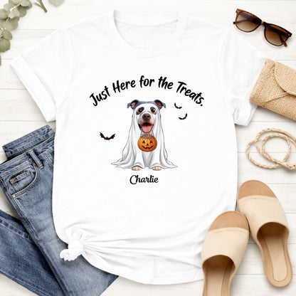 A white graphic T-shirt laid flat on a light wood surface, styled with jeans, sunglasses,...The shirt has a printed dog dressed in a white ghost holding a jack-o'-lantern, with the text 'Just Here for the Treats.' and custom with dog's name printed below
