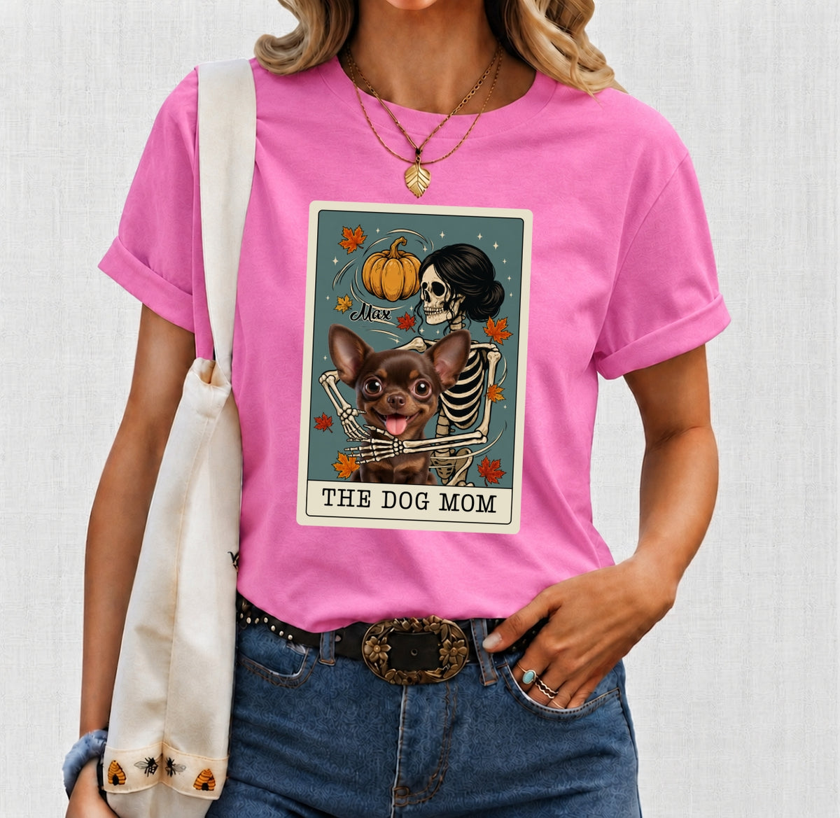 A woman wearing a bright pink graphic T-shirt with an Autumn Halloween-style tarot card design featuring a skeleton and custom dogs