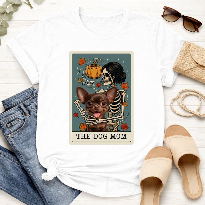 A white short-sleeve T-shirt laid flat on a light wood surface. The shirt features a large rectangular graphic print showing a skeleton hugging a custon dog, with autumn leaves, a pumpkin, and the text 'THE DOG MOM'.