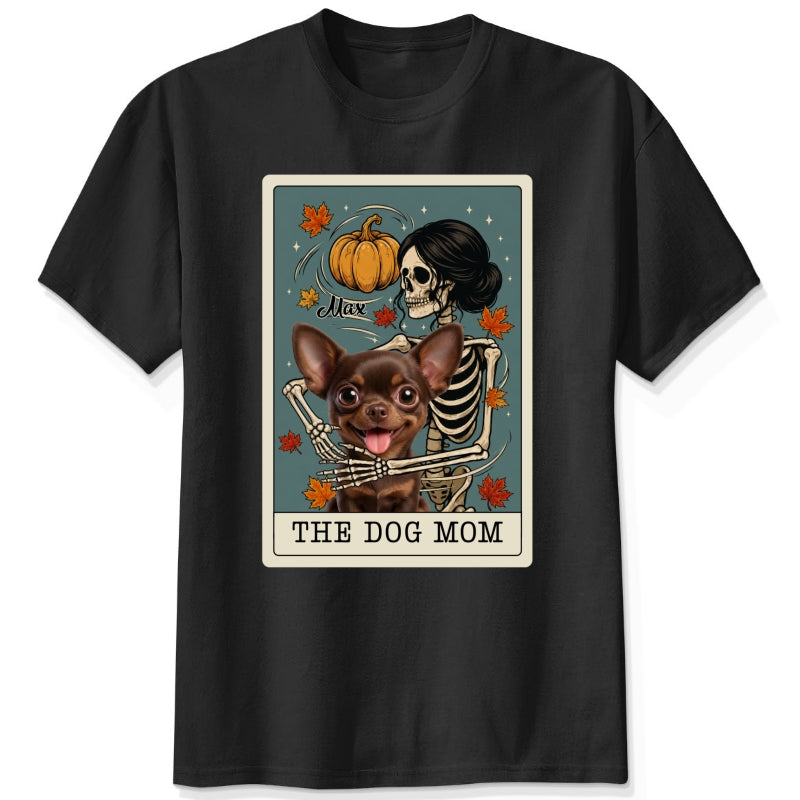 A black short-sleeve crewneck t-shirt featuring a graphic print of a skeleton dog mom and a custom dog under a autumn pumpkin, styled like a tarot card labeled 'THE DOG MOM'.