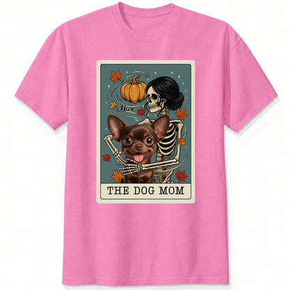 A pink short-sleeve crewneck t-shirt featuring a graphic print of a skeleton dog mom and a custom dog under a autumn pumpkin, styled like a tarot card labeled 'THE DOG MOM'.