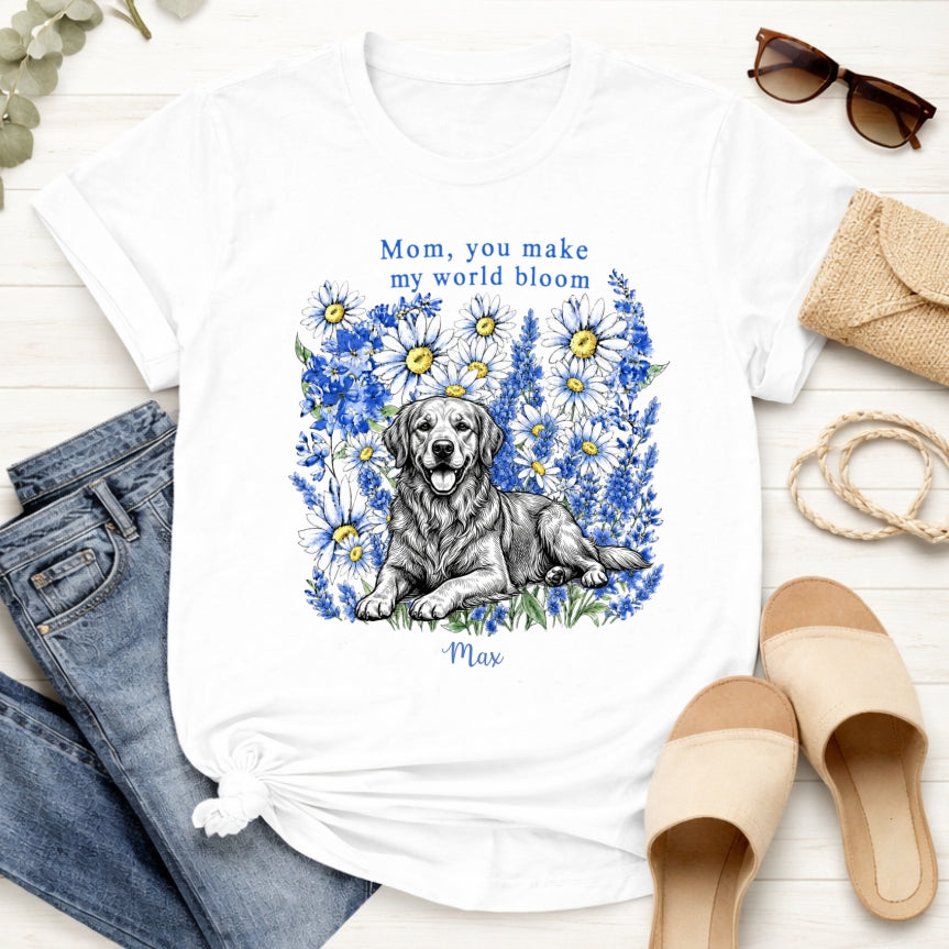 A white custom dogs t-shirt featuring a graphic print of a golden retriever dog surrounded by blue flowers, with the text 'Mom, you make my world bloom' and dog's name written below. The t-shirt is styled in a flat-lay arrangement with blue jeans, beige slide sandals, sunglasses, a burlap clutch, and a rope on a white wood background.