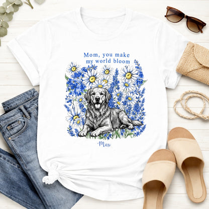 A white custom dogs t-shirt featuring a graphic print of a golden retriever dog surrounded by blue flowers, with the text 'Mom, you make my world bloom' and dog's name written below. The t-shirt is styled in a flat-lay arrangement with blue jeans, beige slide sandals, sunglasses, a burlap clutch, and a rope on a white wood background.