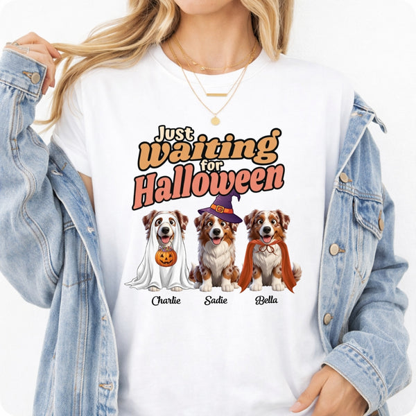 A woman wearing a white graphic T-shirt with a Halloween-themed print featuring three dogs in costumes and the text 'Just waiting for Halloween.' She has a light blue denim jacket off her shoulders against a plain white background.