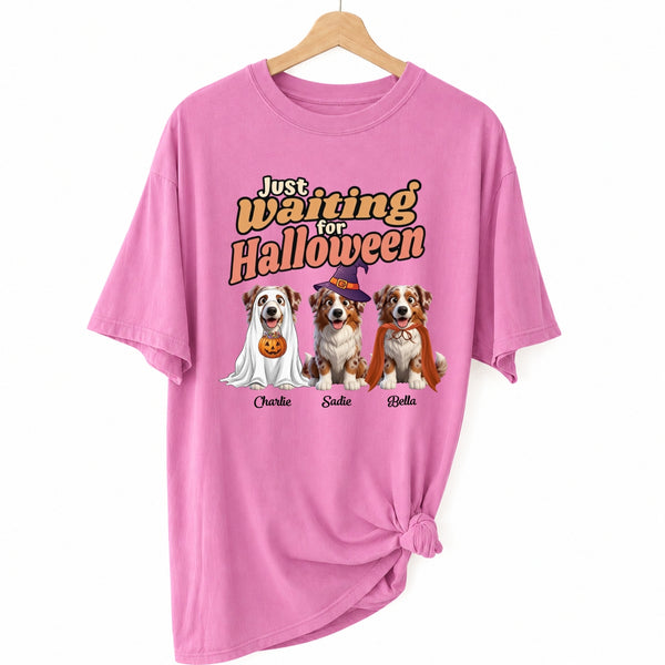 A pink short-sleeve t-shirt hanging on a wooden hanger against a white background, styled with a knot at the bottom right. The shirt features a graphic print of dogs dressed in Halloween costumes with the text 'Just Waiting for Halloween' and customized dog names underneath.